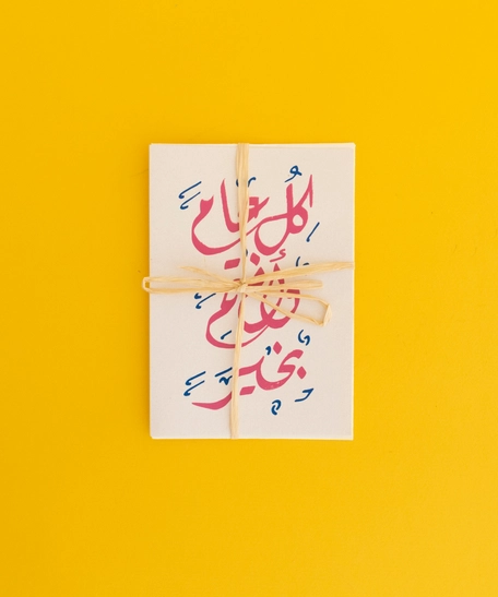 بطاقة معايدة مع ظرف بعبارة "كل عام وأنتم بخير" ومزخرفة بالخط العربي بلون وردي